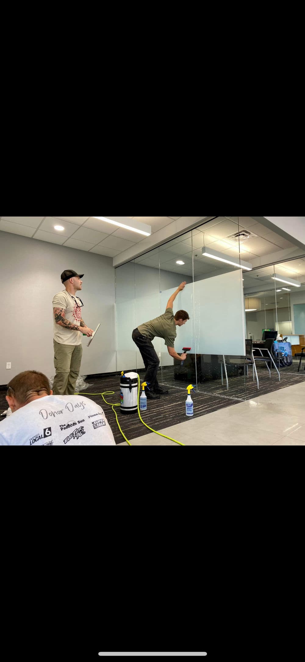 Two workers cleaning glass walls in an office, with cleaning supplies and equipment visible.